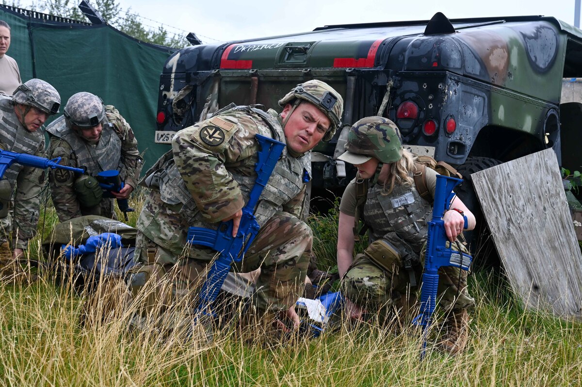 Several airmen wearing camouflage military uniforms with blue training rifles in their hands prepare to lift a simulated casualty off the ground. There is tall grass in the foreground and a military vehicle in the background.