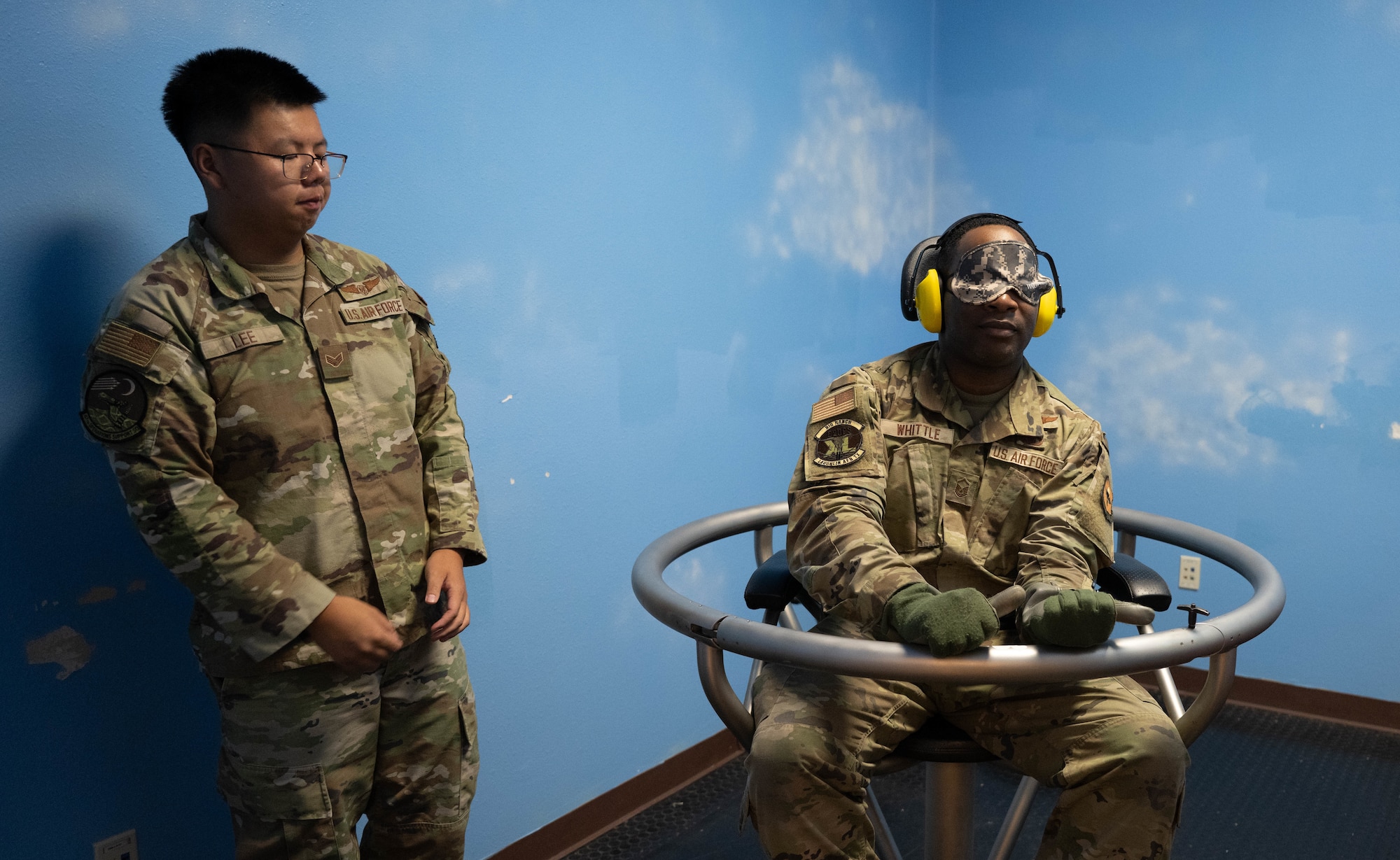 U.S. Air Force Senior Airman Alexander Lee, 47th Operations Support Squadron aerospace physiology technician, observes Master Sgt. Edward Whittle, 47th Flying Training Wing Command Post superintendent, sitting in a Barany Chair at Laughlin Air Force Base, Texas, Aug. 22, 2025.  The Barany Chair was designed for pilots to train for spatial disorientation by accelerating the inner ear. (U.S. Air Force by Airman 1st Class Darryl Keith.)