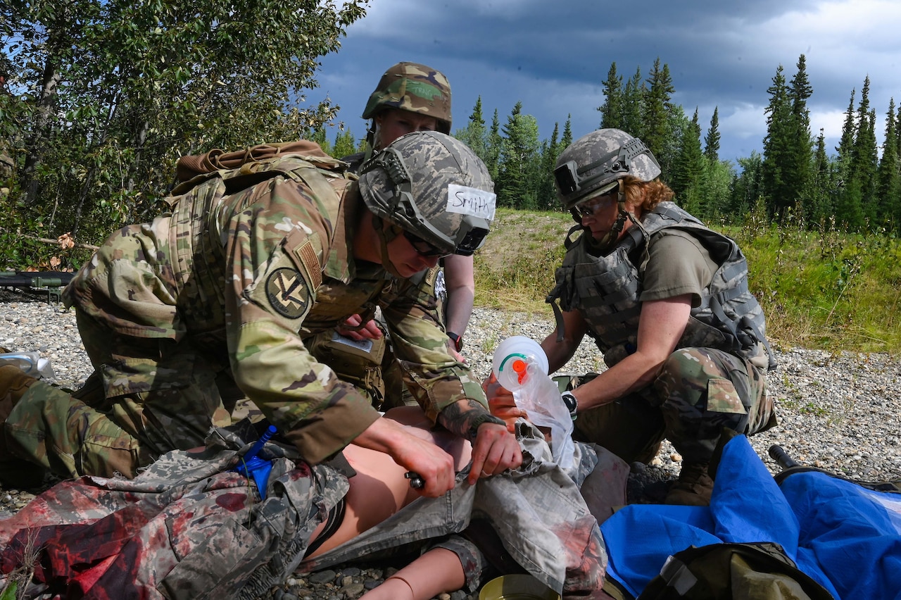 A man wearing a military camouflage uniform and helmet places a needle in a simulated casualty mannequin while a woman in similar attire places an oxygen mask on the mannequin.