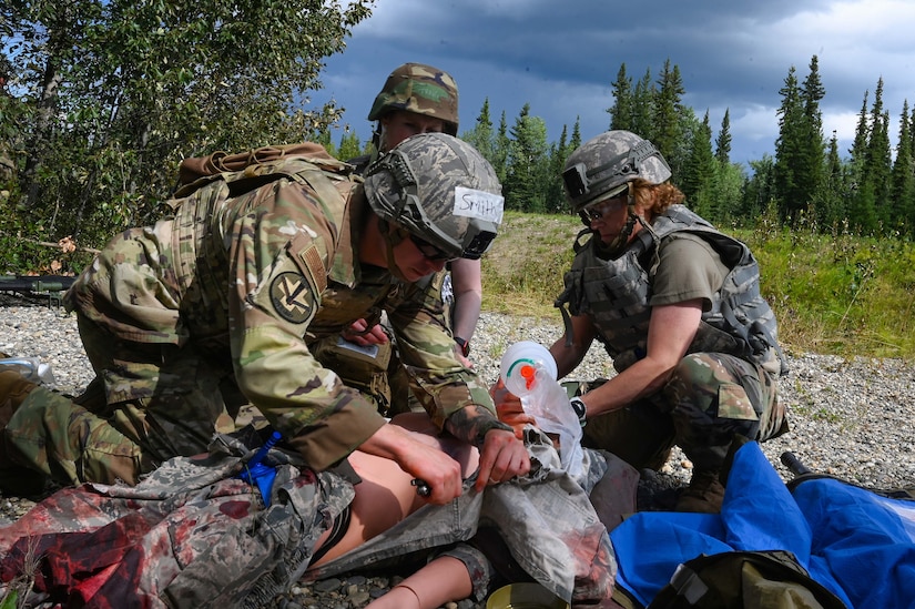 A man wearing a military camouflage uniform and helmet places a needle in a simulated casualty mannequin while a woman in similar attire places an oxygen mask on the mannequin.