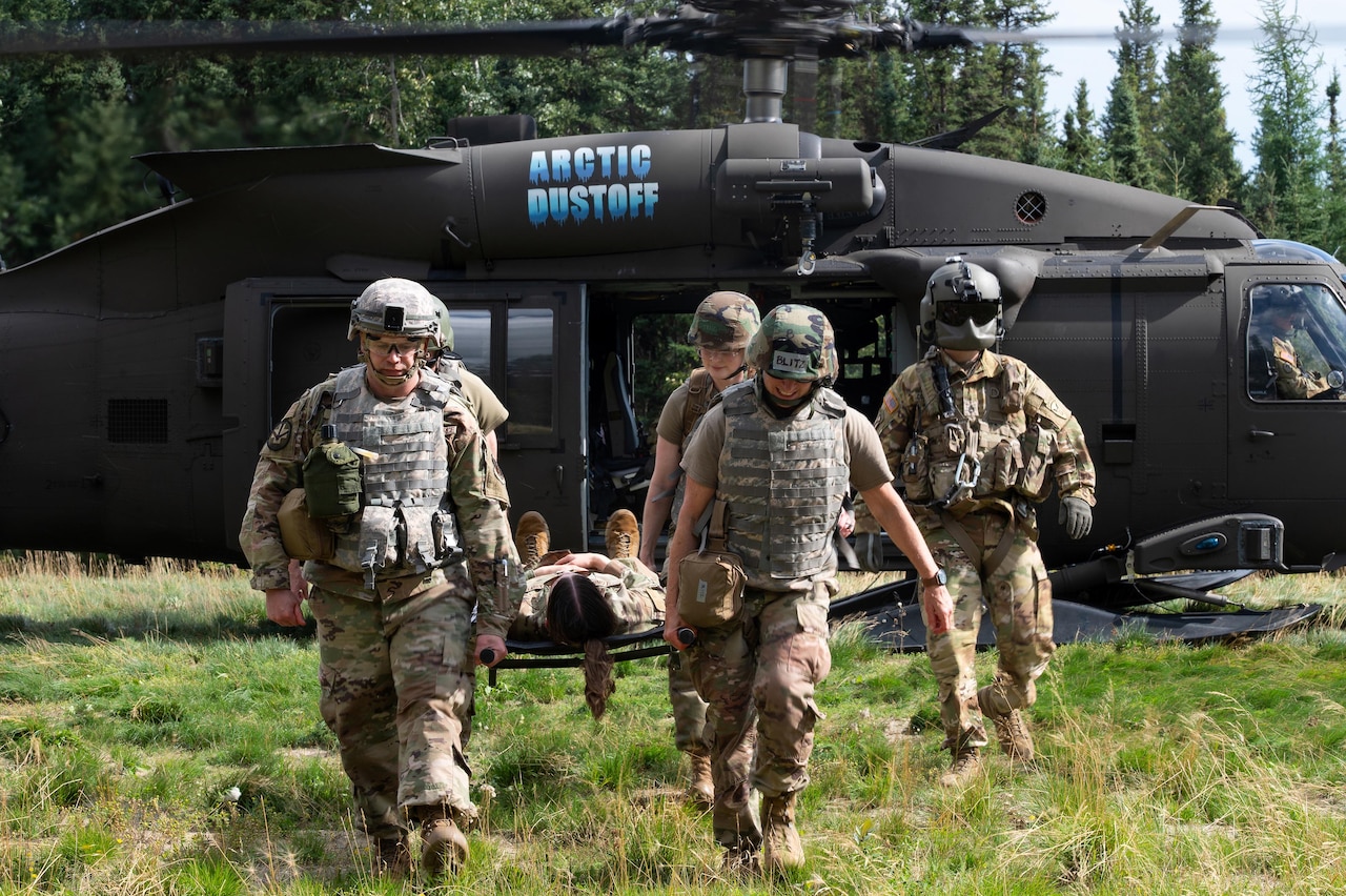 Four airmen wearing camouflage military uniforms, helmets and vests carry a stretcher with another person in a camouflage military uniform on top of it. There is another person wearing a camouflage military uniform and an aviation helmet to the right of the people carrying the stretcher and a helicopter in the background.