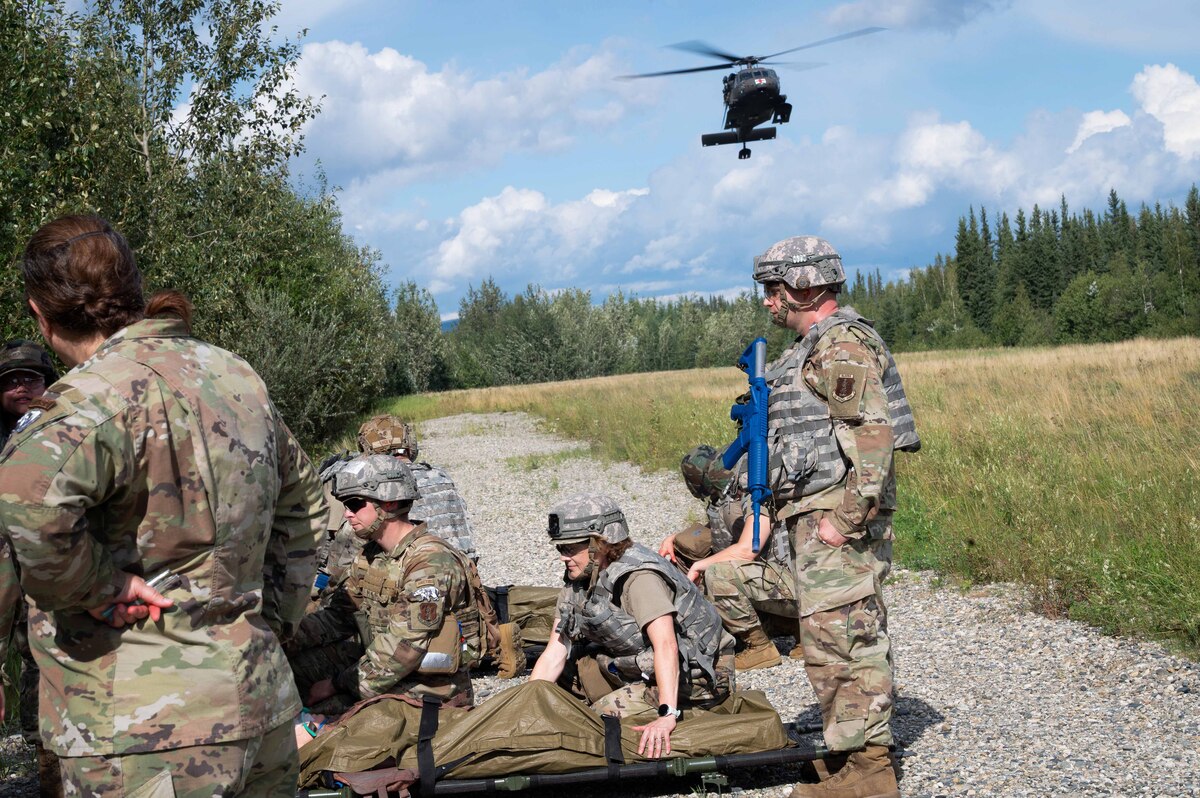Men and women wearing military camouflage uniforms stand and kneel over two stretchers with mannequins inside on a paved road near an open field surrounded by trees. A helicopter is flying above.
