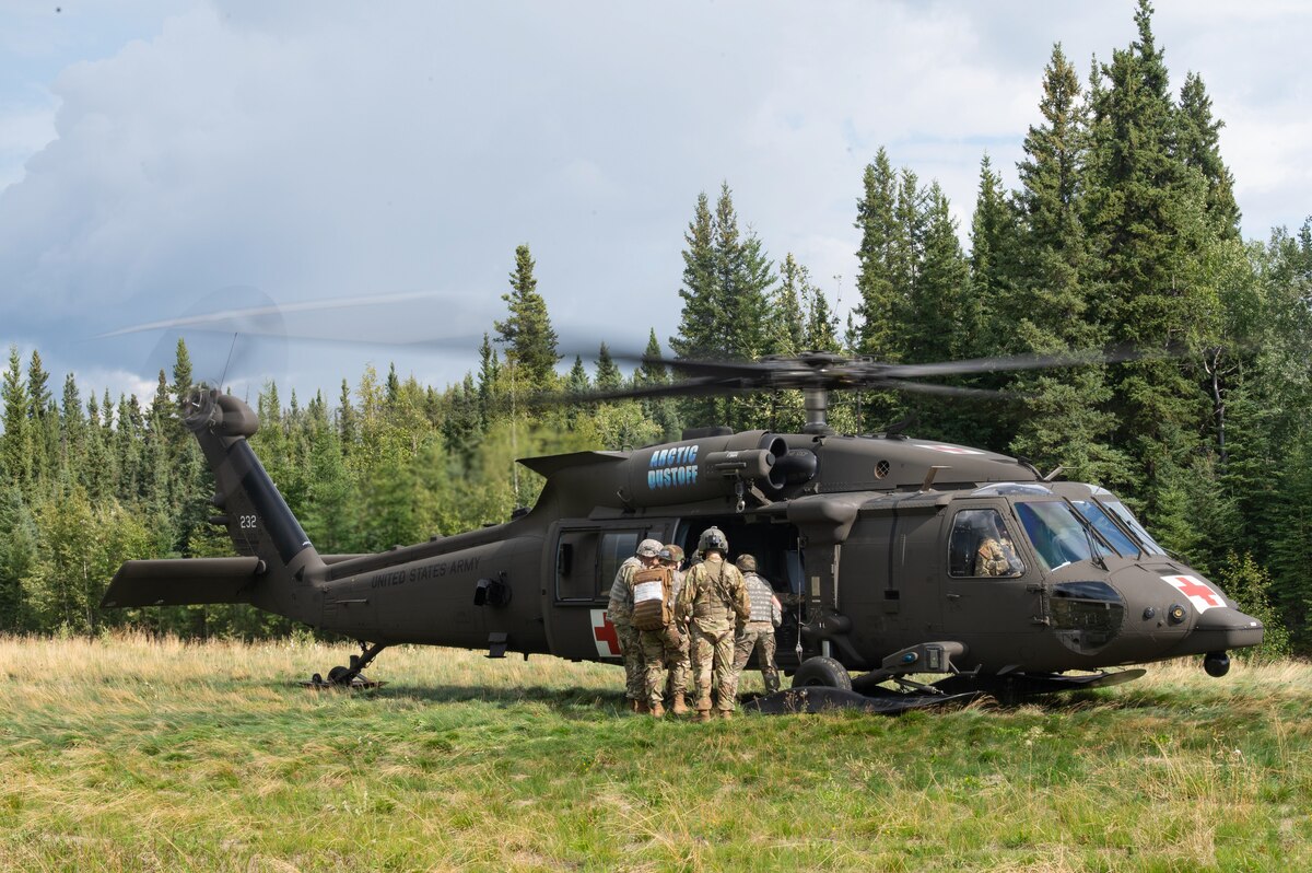 Several airmen wearing camouflage military uniforms load a simulated casualty into a helicopter. The helicopter’s blades are spinning as the pilot waits to takeoff from a field with trees in the background.