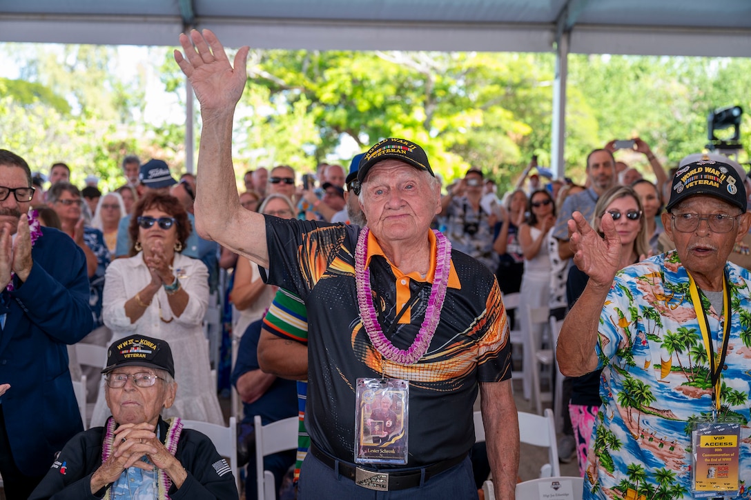 A veteran in a lei and baseball cap waves while standing with other vets in an audience of people clapping at an outdoor event.