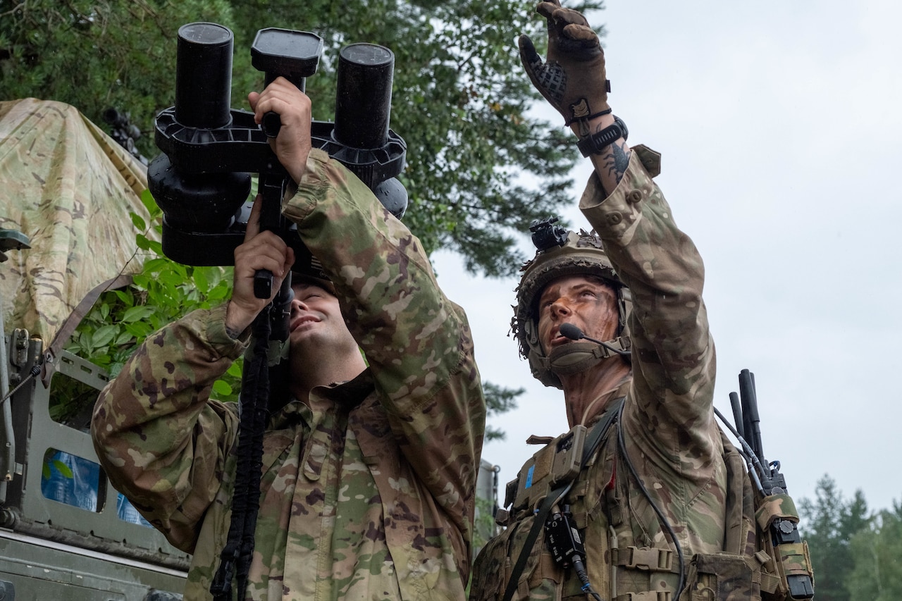 A soldier in a camouflage military uniform points an electronic jamming device toward the sky. Another soldier, standing in similar attire, points at the sky.