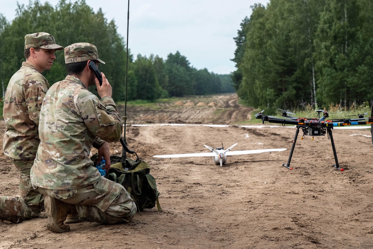 Two soldiers in military camouflage uniforms kneel on a dirt path in a wooded area, with two drones on the ground nearby. The soldier on the right has a backpack in front of him and a phone receiver in his hand.