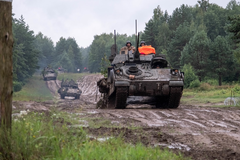 Military tanks roll down a dirt road in a wooded area with soldiers in camouflage military uniforms perched atop of the tanks.