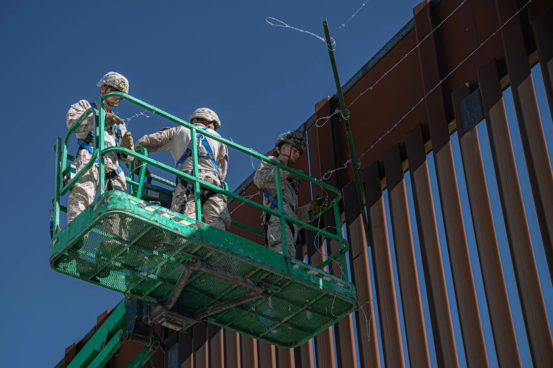 Three Marines stand on a green platform next to a metal wall under blue skies.