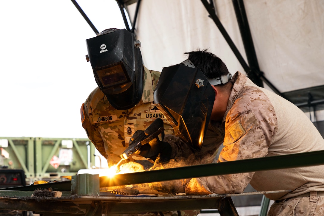 A soldier in a helmet watches another solider weld two metal pieces together inside a large tent structure.