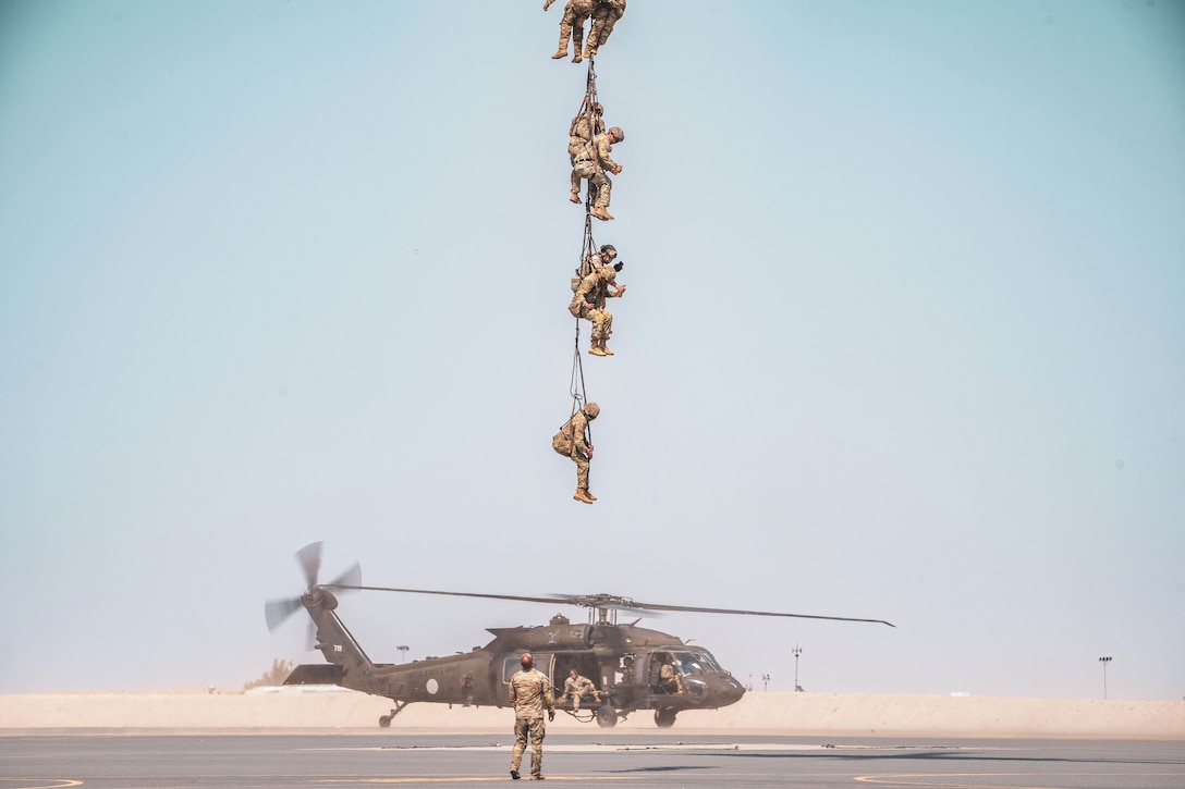 Seven soldiers connected by rope hover in a hazy sky as one soldier watches from below and others sit in parked helicopter.