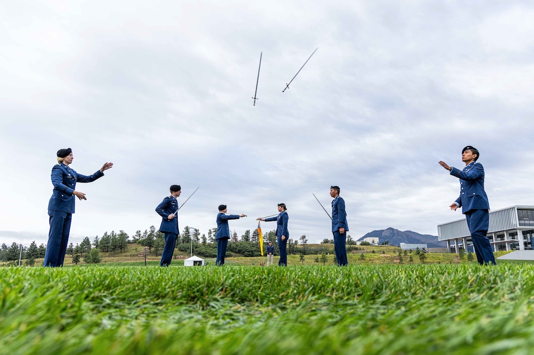 U.S. Air Force Academy cadets toss swords while standing in formation in a field under a cloudy sky with mountains in the background.