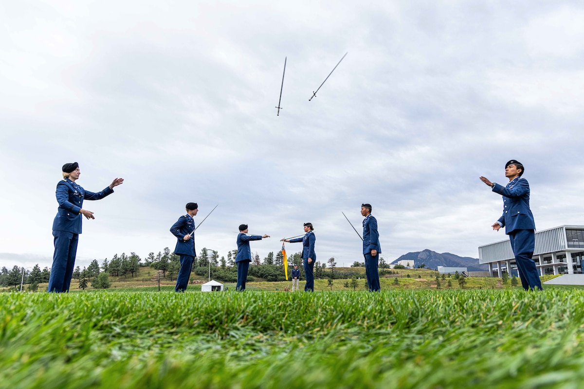 U.S. Air Force Academy cadets toss swords while standing in formation in a field under a cloudy sky with mountains in the background.