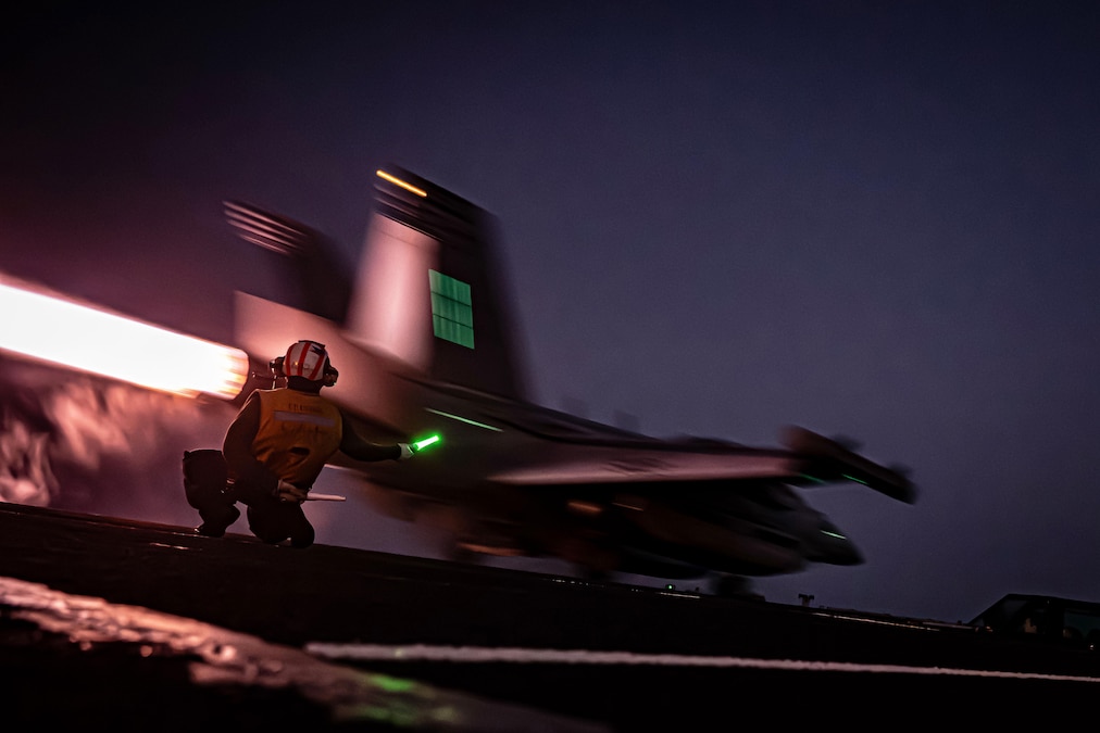 A sailor with a baton directs an aircraft in the dark as it launches from a ship illuminated by white lights.