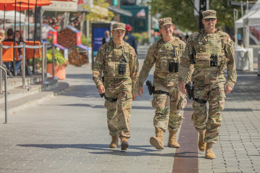 Three guardsmen walk next to each other down a sidewalk in between buildings and outdoor structures on a sunny day.