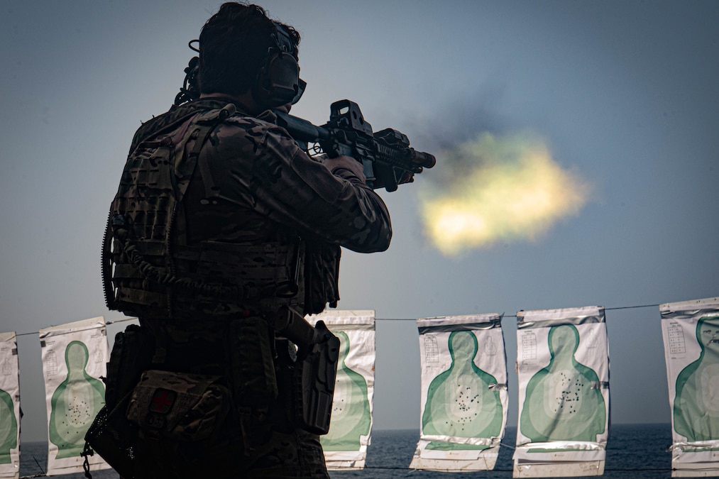 A sailor in tactical gear fires a service weapon at a row of hanging targets with the ocean in the background, as seen from behind.