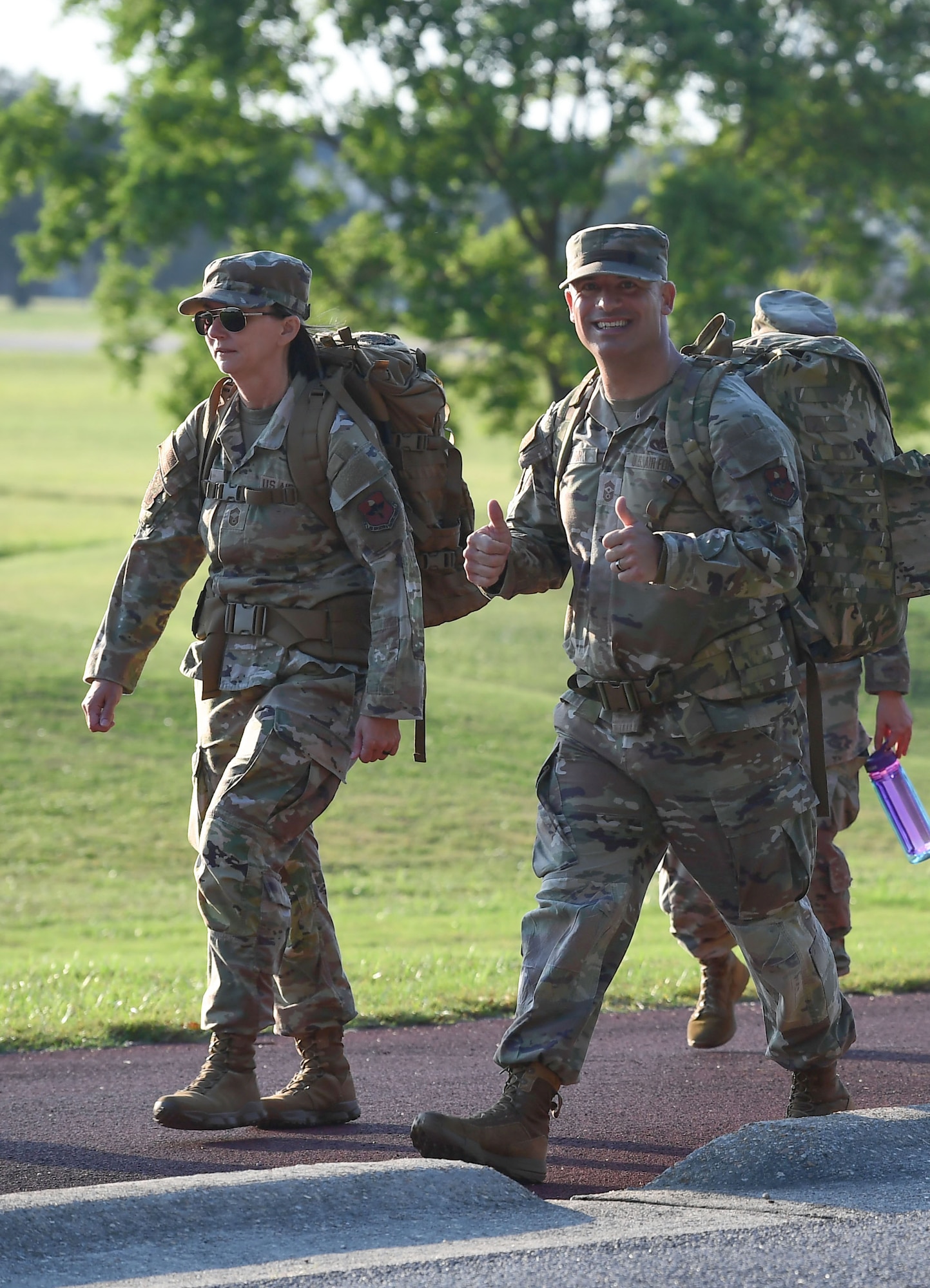 A man and a woman in military uniform march while wearing ruck sacks on their backs.