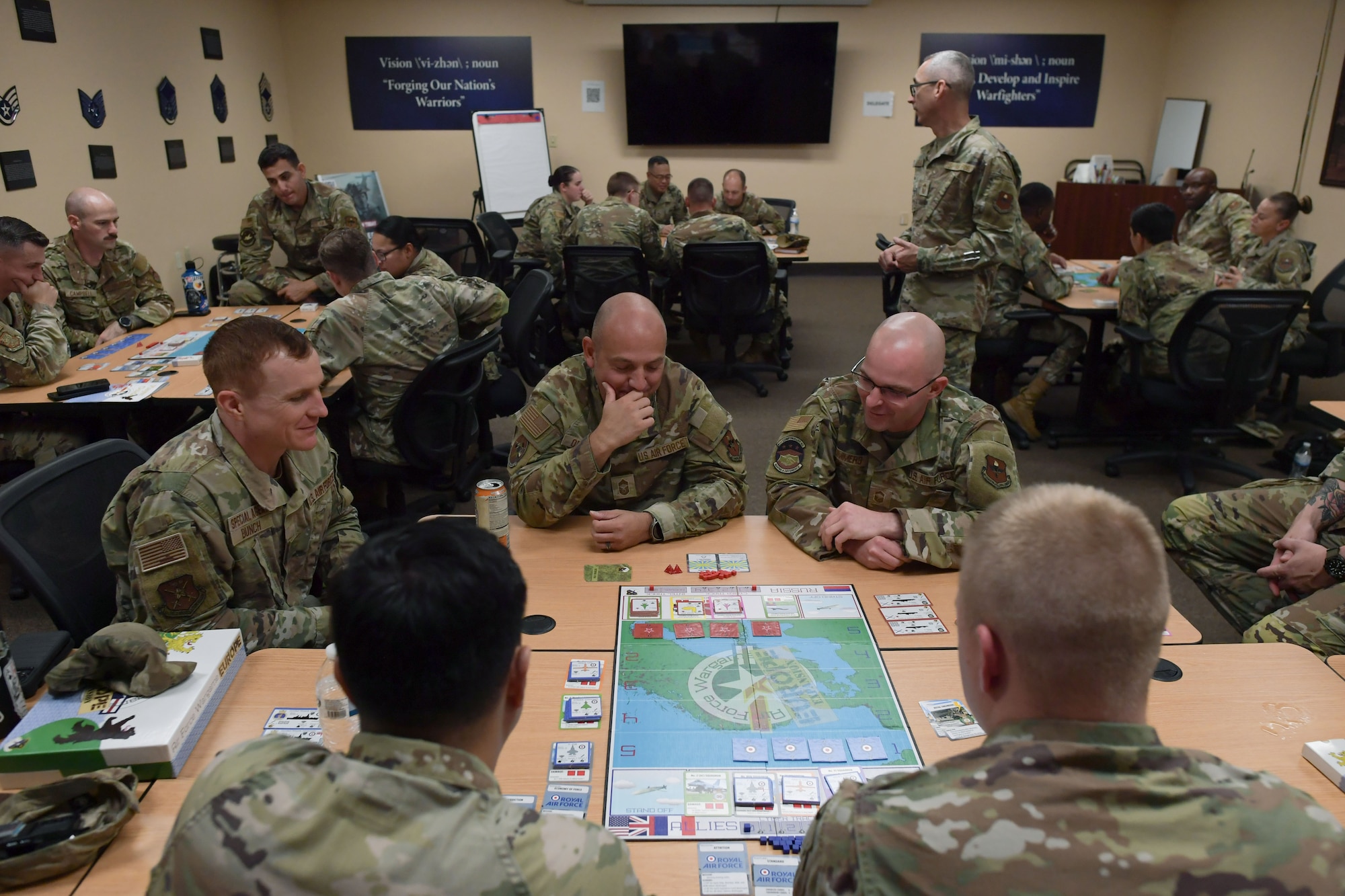 A group of people in military uniform sit around tables playing a board game.