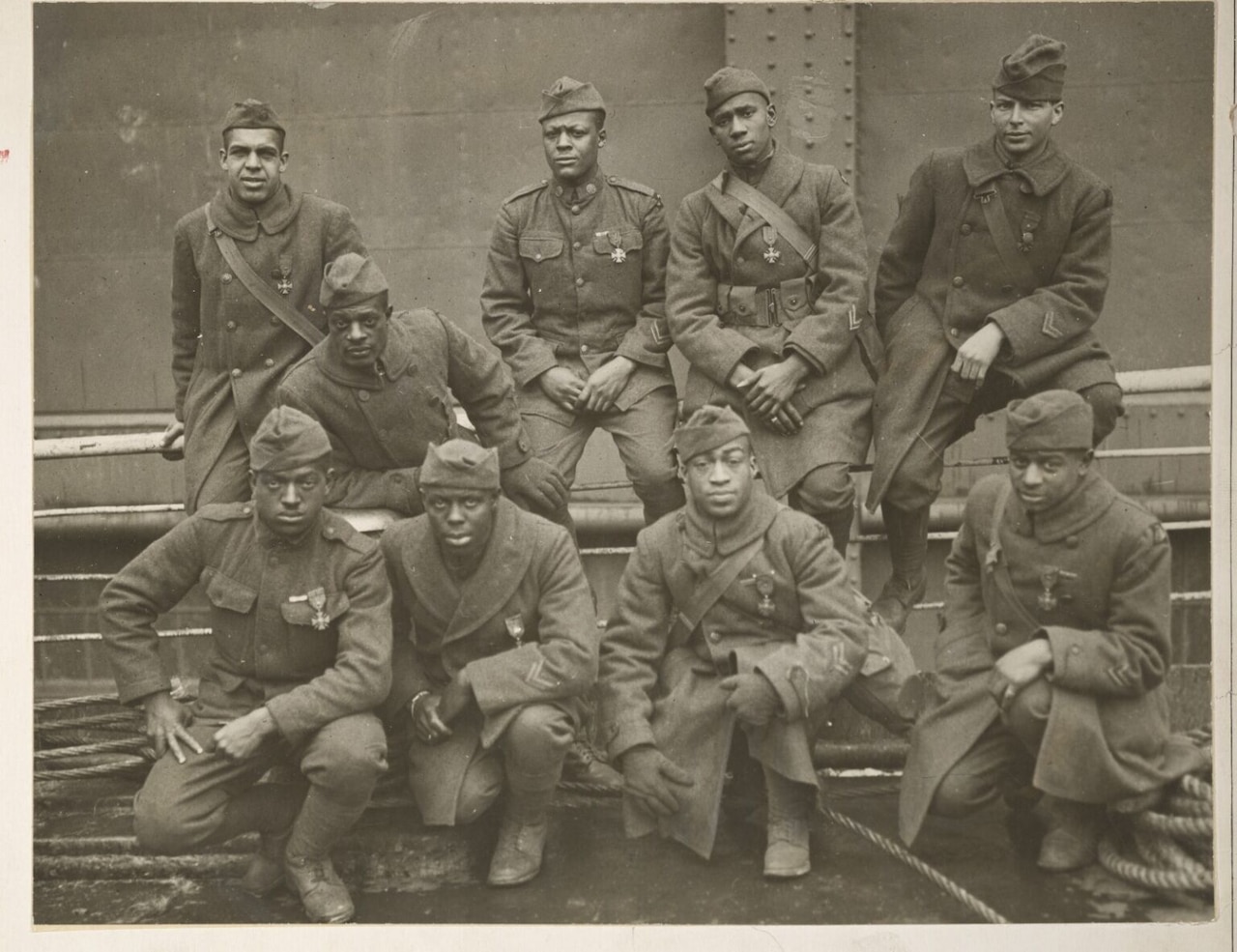 A group of nine men in early 20th-century military uniforms are posing for a photograph. Four of the men are sitting on fencing in the background, and four are kneeling in the foreground, with the 9th man sitting between both groups.
