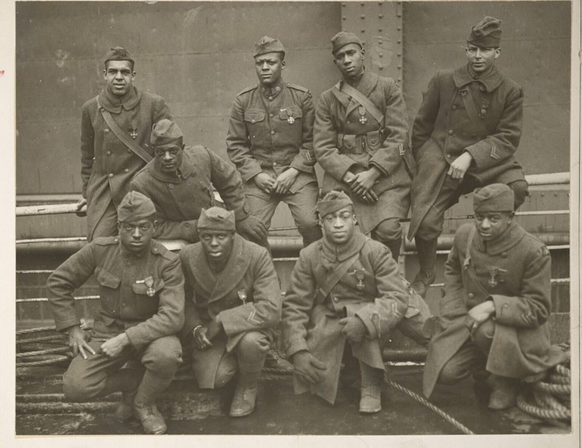 A group of nine men in early 20th-century military uniforms are posing for a photograph. Four of the men are sitting on fencing in the background, and four are kneeling in the foreground, with the 9th man sitting between both groups.