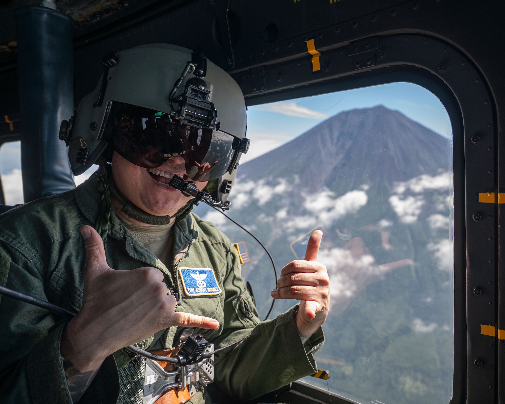 A servicemember smiles in a helicopter flying in front of a mountain