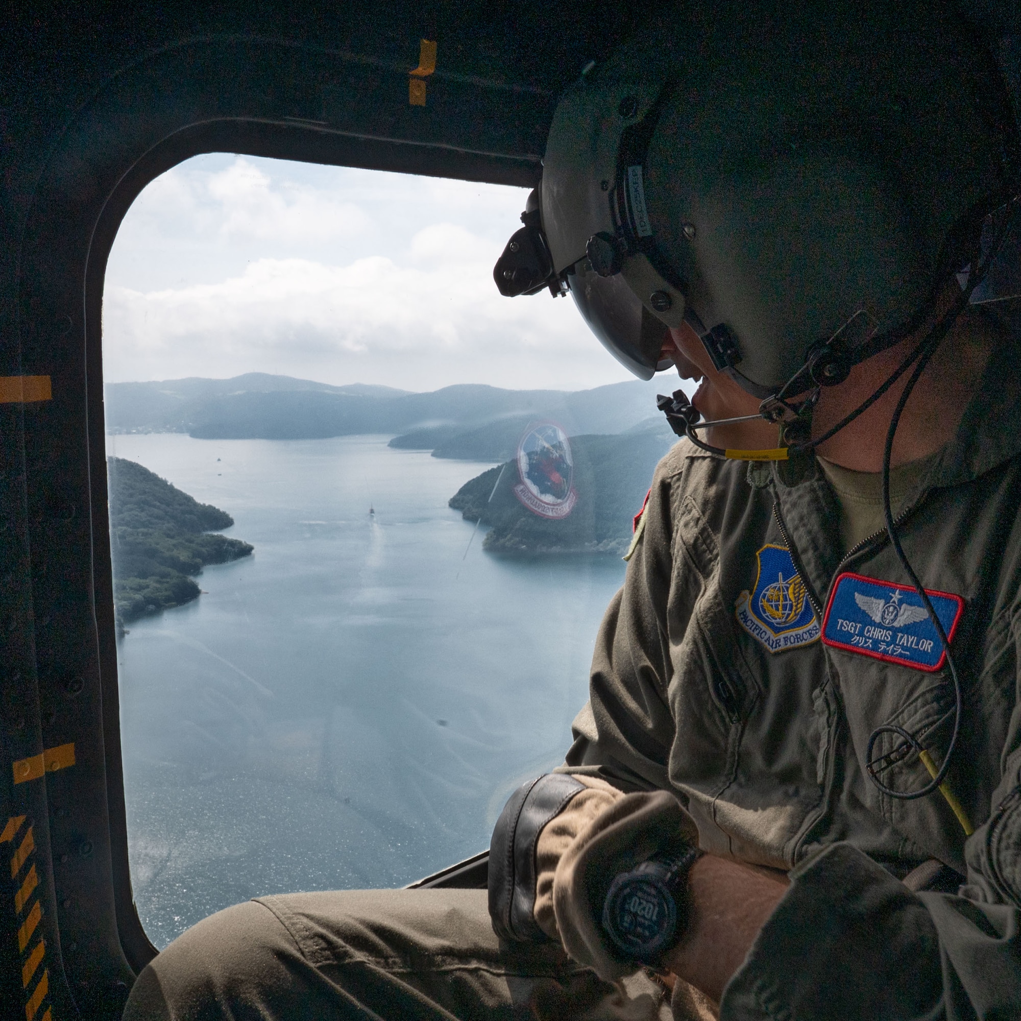 A servicemember looks out of a helicopter to the coast