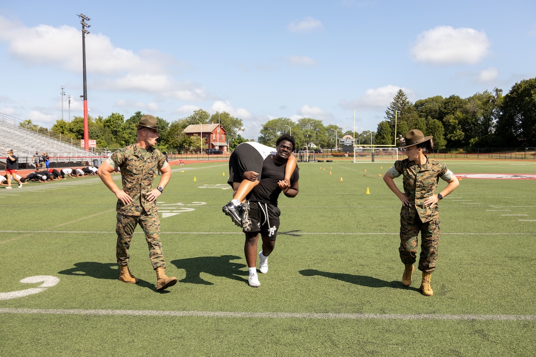 U.S. Marine Corps Sgt. Elizabeth McSparin, right, a drill instructor and Marine Corps Instructor of Water Survival with Bravo Company, 1st Recruit Training Battalion, Marine Corps Recruit Depot Parris Island, and Staff Sgt. Ean Effert, left, a drill instructor with Golf Company, 2nd Recruit Training Battalion, Marine Corps Recruit Depot Parris Island, motivates a Brockton High School football player during a Marine Corps combat fitness test as part of Marine Week Boston, Brockton, Massachusetts, Aug. 25, 2025. During Marine Week Boston, Marines with Marine Corps Recruiting Command shared Marine Corps traditions with local high schools in honor of the Corps’ 250th birthday. Marines stationed across the globe commemorate the 250th anniversary of the Marine Corps, honoring a distinguished legacy of service, sacrifice, and unwavering fidelity to both the nation and fellow Marines. (U.S. Marine Corps photo by Staff Sgt. Erin A. VandeHoef)