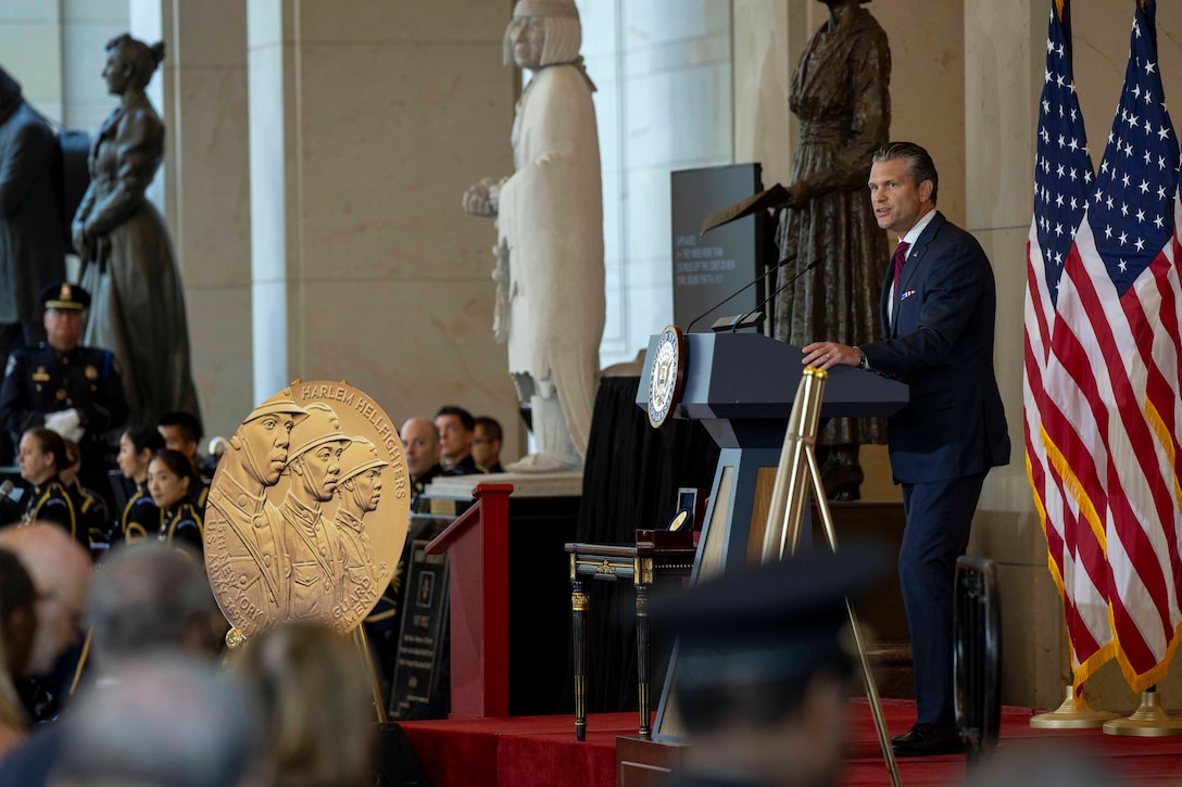 A person in a business suit stands and speaks at a lectern, with two American flags behind the lectern, and a gold symbol with three faces on it also displayed; seated people are in the audience.