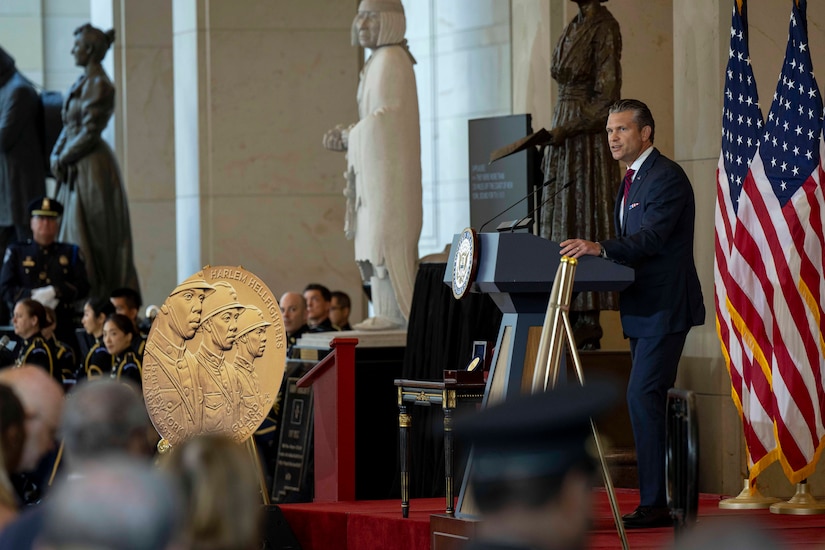 A person in a business suit stands and speaks at a lectern, with two American flags behind the lectern, and a gold symbol with three faces on it also displayed; seated people are in the audience.