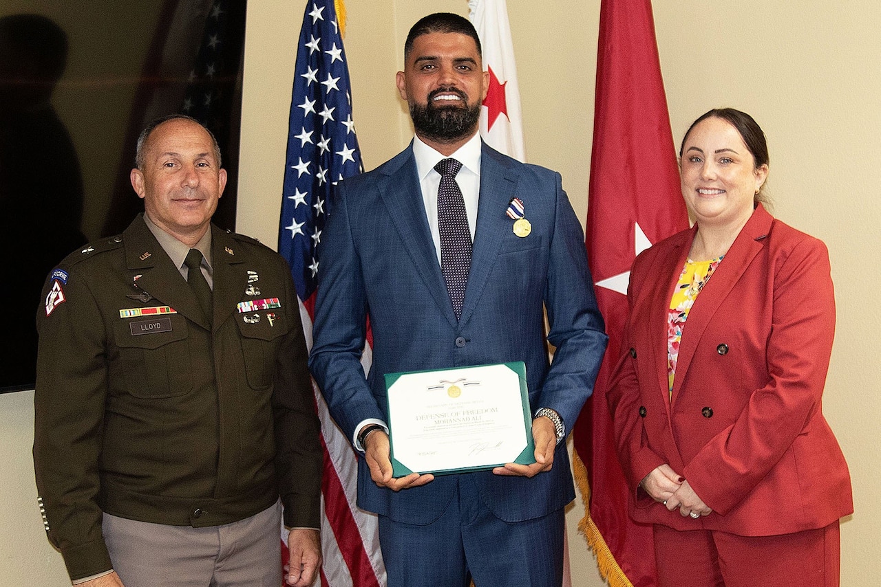 A man, center, wearing civilian attire holds a certificate and smiles. There is a man in a military dress unform standing to the left and a woman in civilian attire standing to the right. There are three flags in the background, the flag to the left us the American flag.