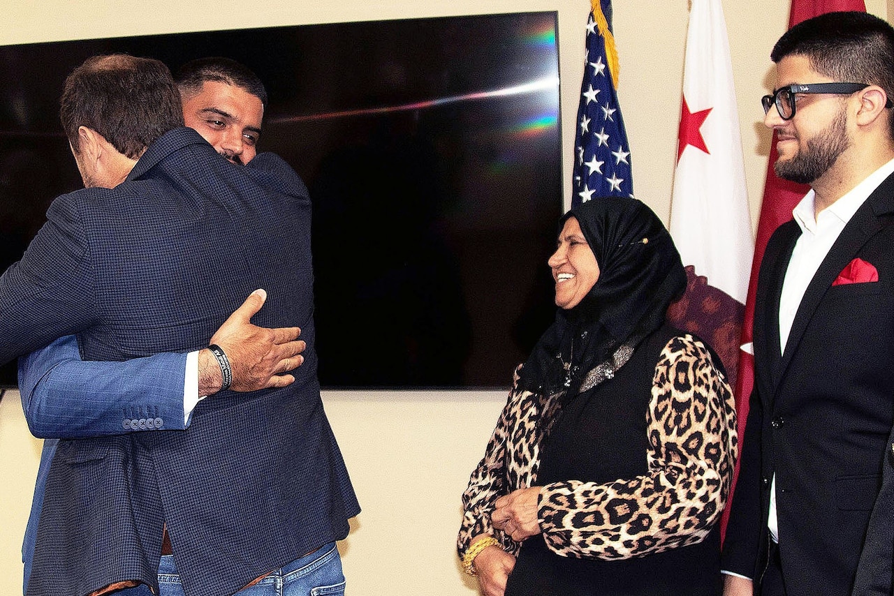 Two men dressed in civilian attire are hugging while a woman dressed in hijab and a man wearing civilian attire watch. In the background there are flags, including the American flag.