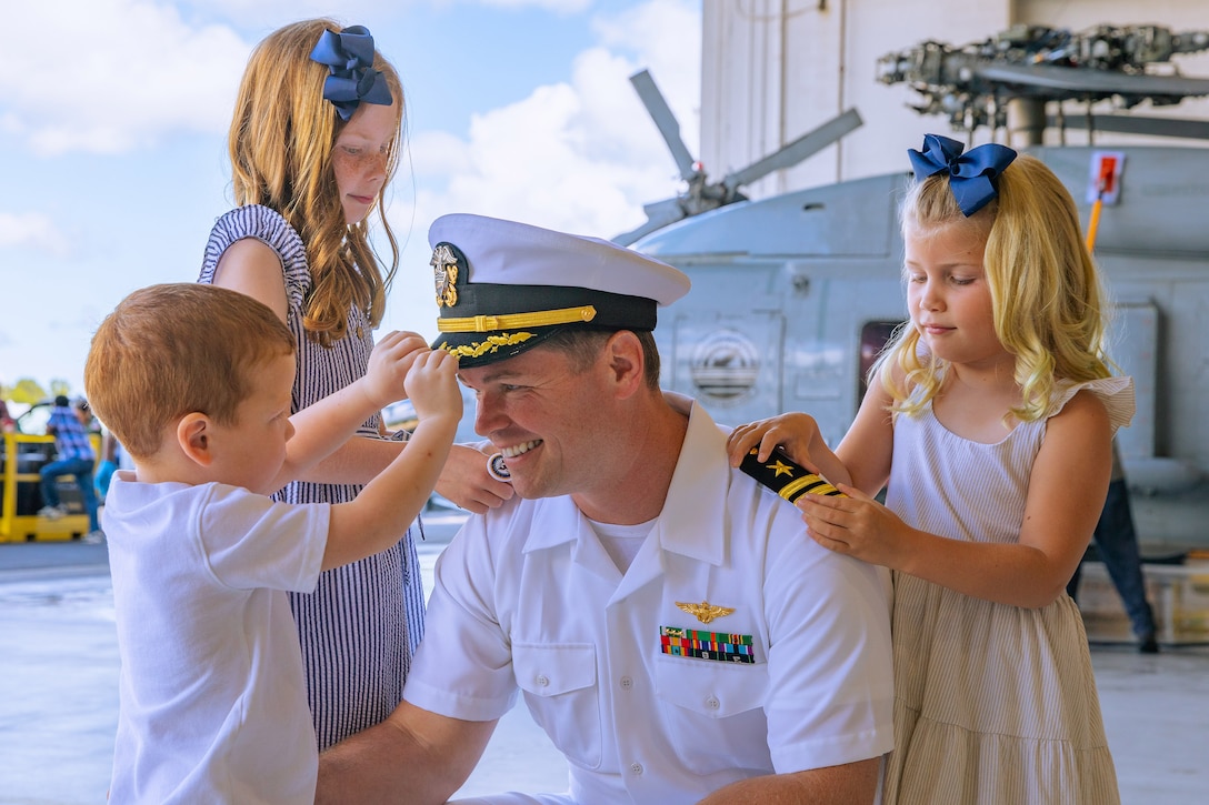 A smiling sailor squats as three kids surround them in a hangar bay with a helicopter in the background.