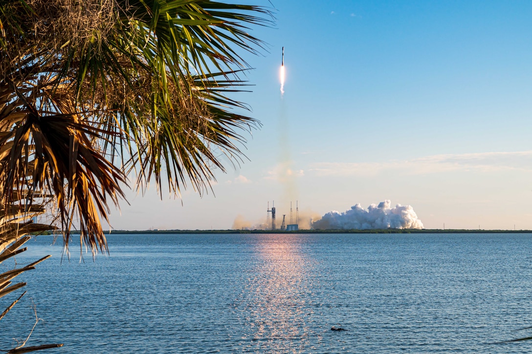 A rocket launches into a blue sky leaving behind clouds of smoke in front of a body of water with a partial palm tree visible on the left.