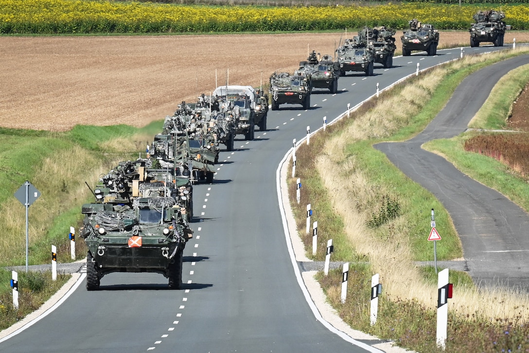 More than a dozen military vehicles drive in formation on a road during the day surrounded by fields.