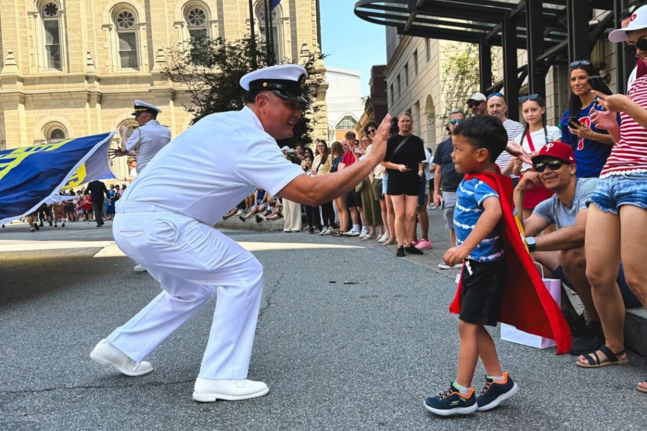 A sailor in dress military uniform gives a high-five to a little boy wearing a red cape during a parade.