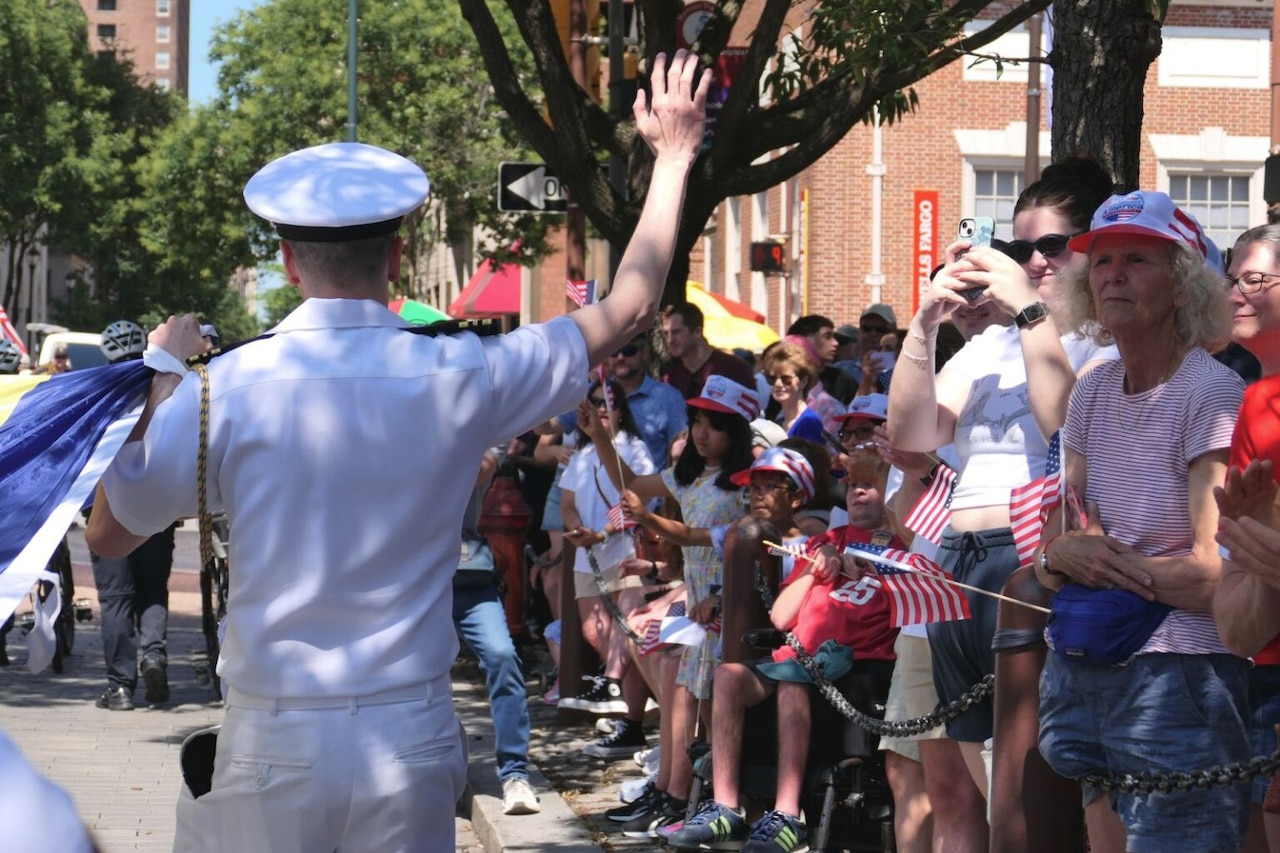 A sailor wearing a dress military uniform waves to a crowd of people at a parade. The people are dressed in casual attire, and many are holding small American flags.