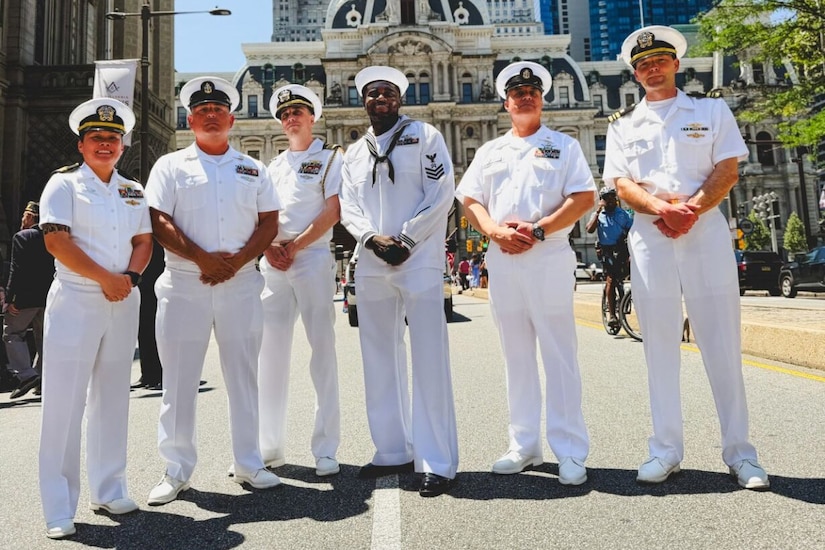 A group of sailors in dress military uniforms pose for a photo with large buildings in the background.