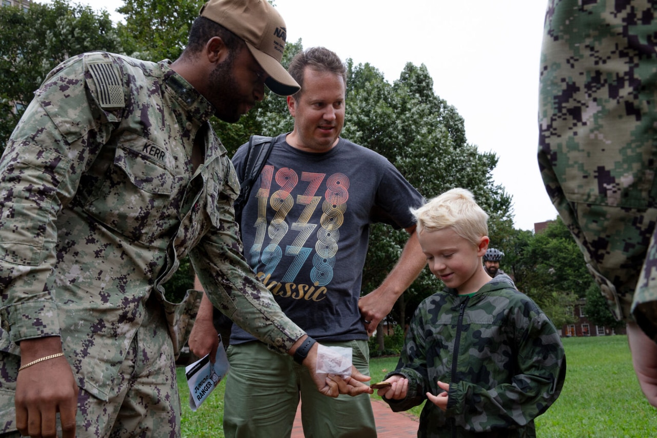 A sailor wearing a military camouflage uniform hands a child a Navy coin. A man in casual attire is standing next to the boy.