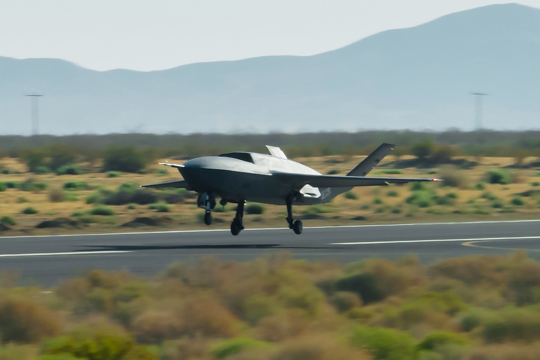 An aircraft takes off from a runway in a sparsely vegetated area, with a mountain range in the background.
