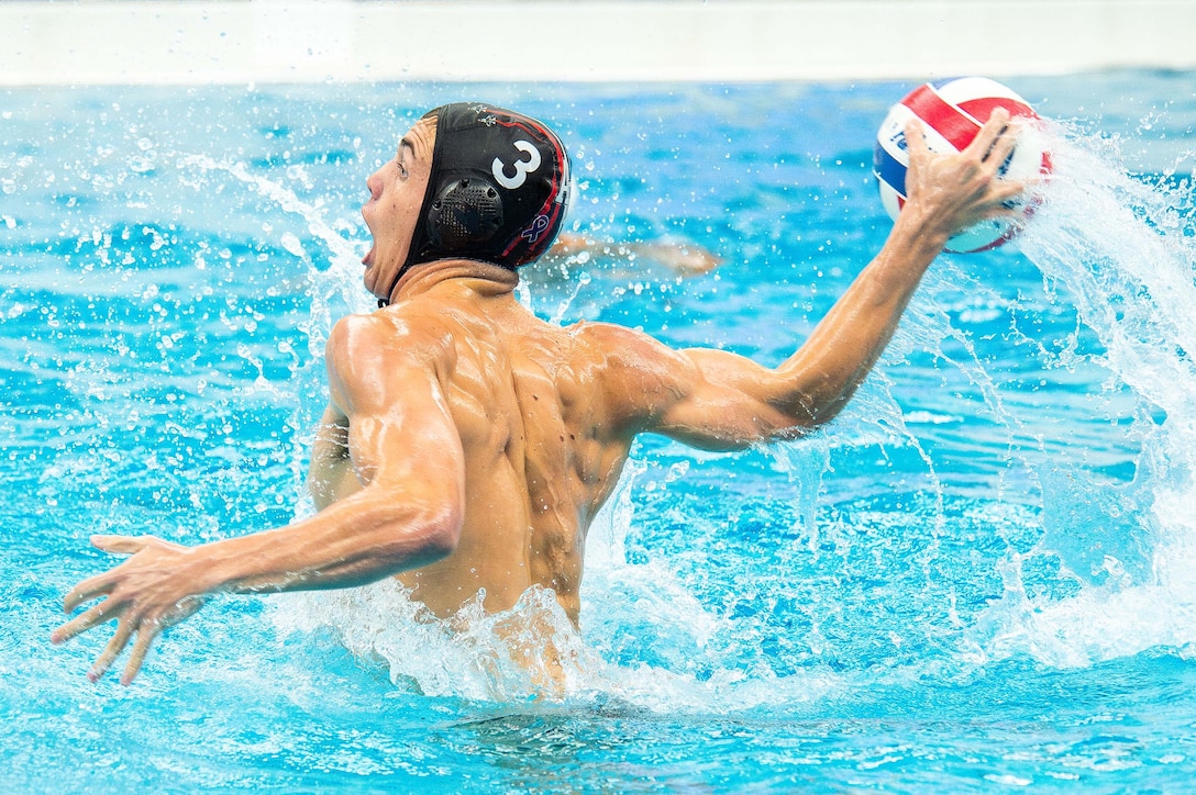 A person in a pool with a black swimming cap on holds a red and blue volleyball in their right hand, preparing to throw, as seen from behind.