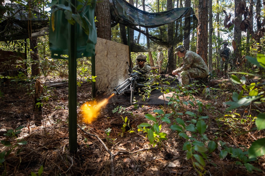 A soldier in a helmet fires a machine gun in a forest setting next to a wooden board while another soldier sits nearby and watches.