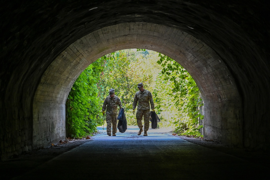Two guardsmen each carry a black trash bag and walk through a tunnel with a sunlit forest setting behind.