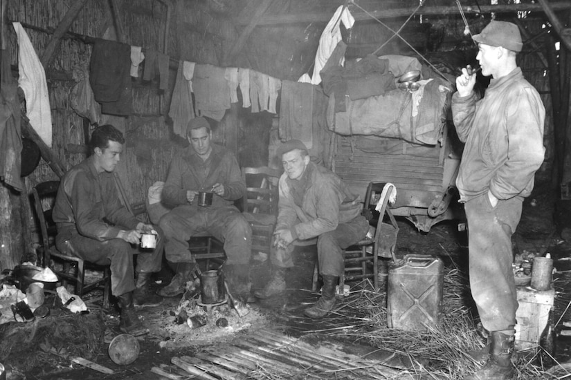 Three men sit in chairs in a shed filled with gadgets. A fourth man stands, smoking a pipe.