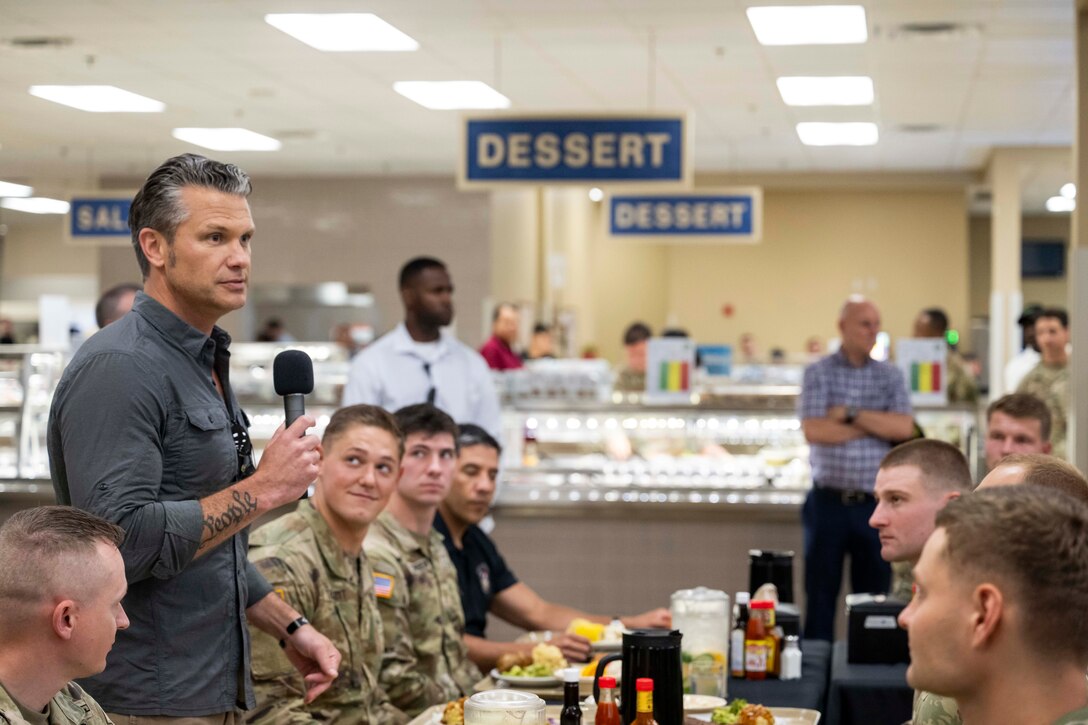 Defense Secretary Pete Hegseth stands and holds a mic while speaking to soldiers seated around him in a dining facility with signs and other people in the background.