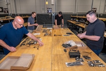 Four men unboxing and organizing small mechanical parts at a work table.