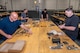 Four men unboxing and organizing small mechanical parts at a work table.