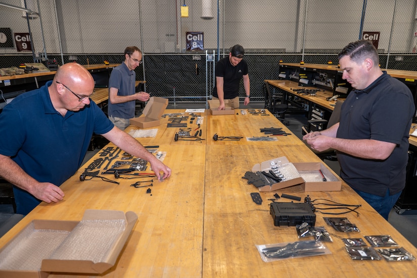 Four men unboxing and organizing small mechanical parts at a work table.