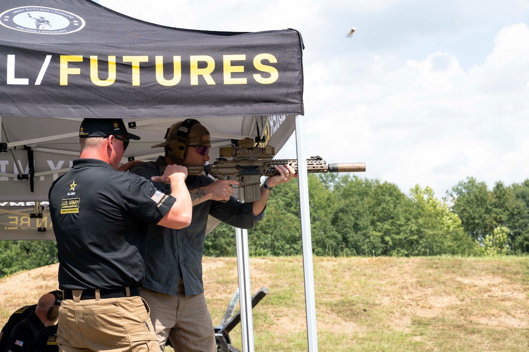 Defense Secretary Pete Hegseth fires a Next Generation Squad Weapon system with an instructor under an outdoor tent.