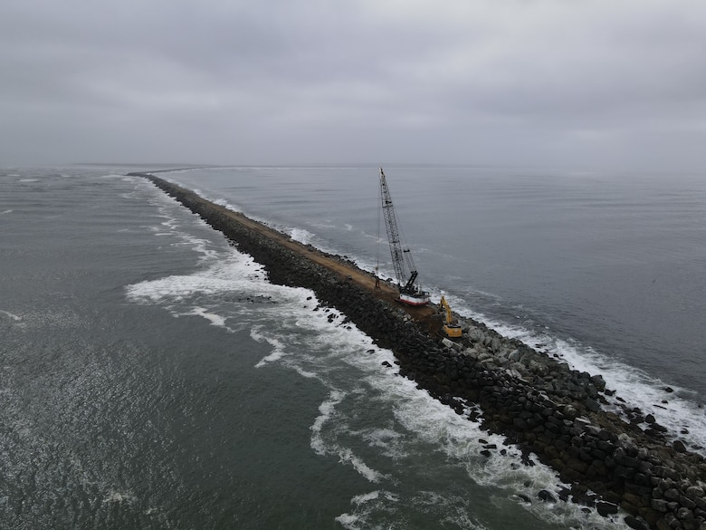 A rock structure coming out of the mouth of a river is pictured with a crane and a bulldozer on top.
