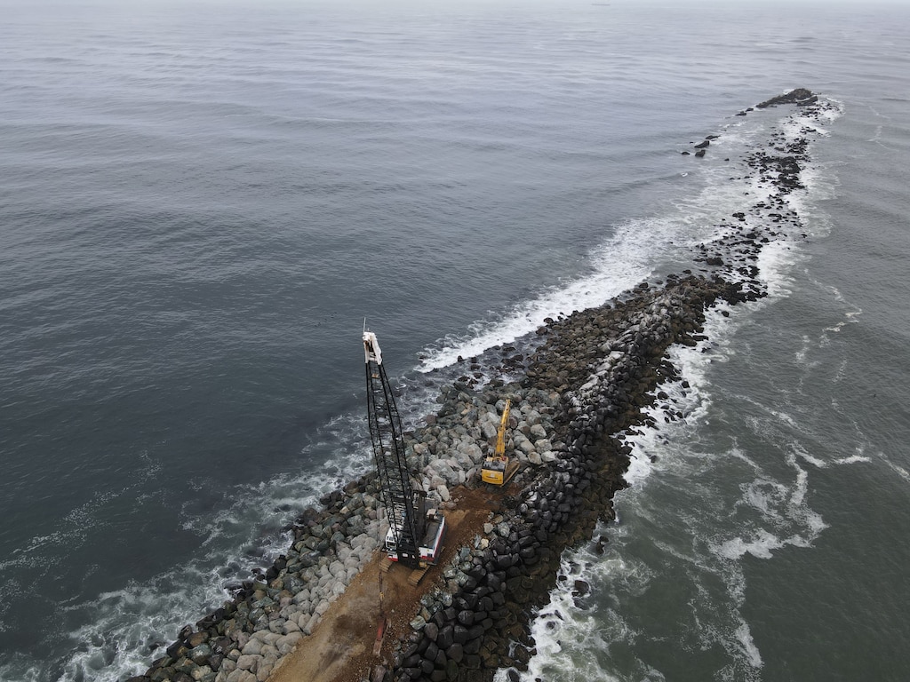 A rock structure coming out of the mouth of a river is pictured with a crane and a bulldozer on top.