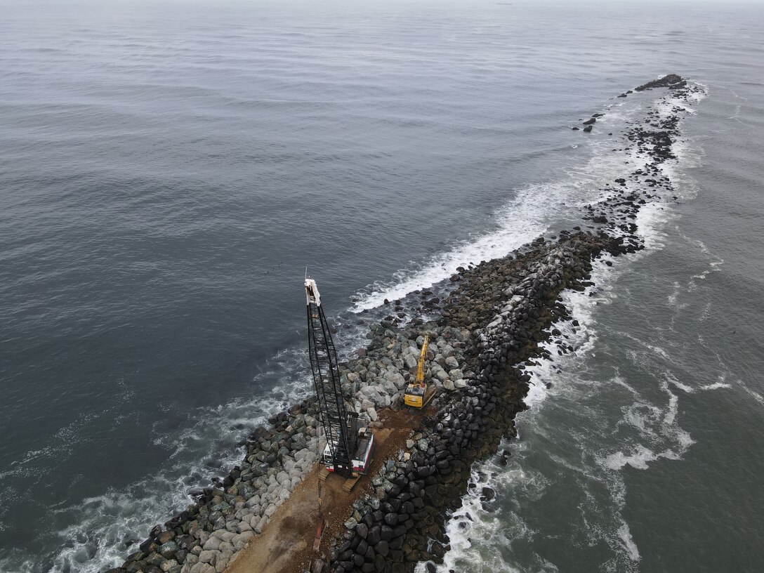 A rock structure coming out of the mouth of a river is pictured with a crane and a bulldozer on top.
